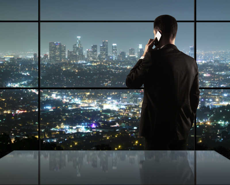 夜景下的站在窗边打电话看着城市风景的商人