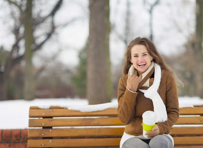 冬日里坐在长椅上拿着咖啡杯的微笑的年轻女子