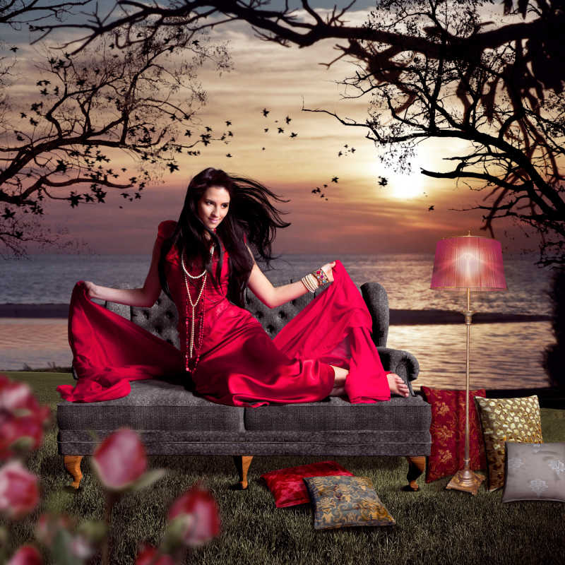 漂亮的女人穿着红色丝绸晚礼服坐在湖边的旧沙发上