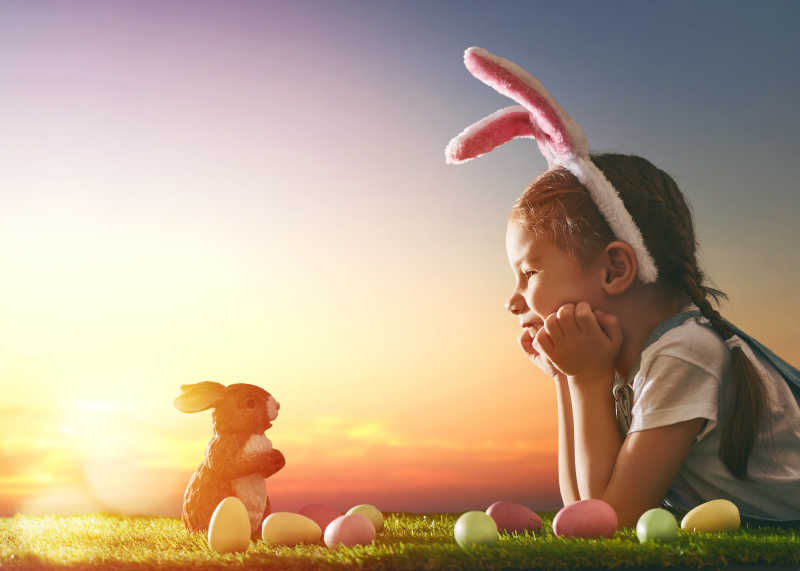 在复活节戴兔子耳朵的小女孩趴在草地上旁边还有草地和兔子
