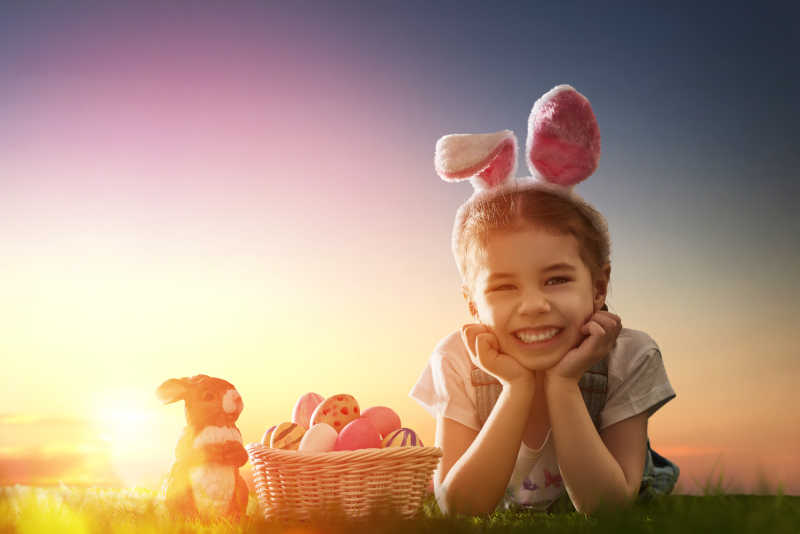 趴在草地上戴着兔子发箍的快乐的小女孩旁边复活节彩蛋兔子