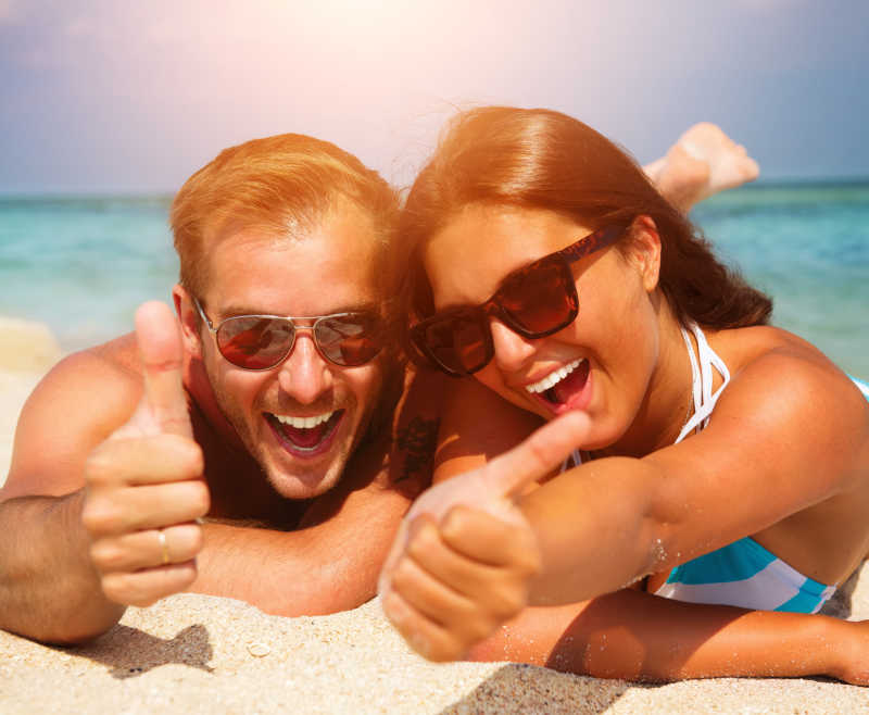 幸福的夫妇戴着太阳镜趴在沙滩上竖起大拇指