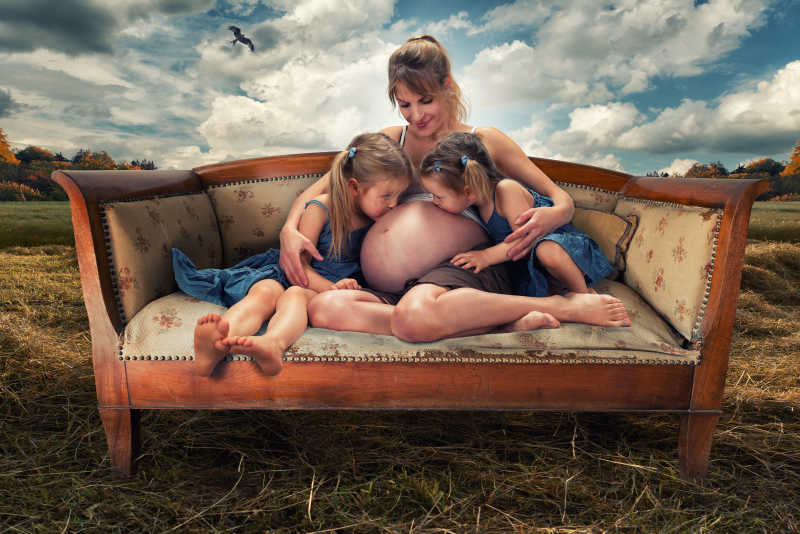怀孕的母亲抱着两个小女孩坐在大自然的长凳上