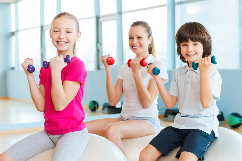 健康俱乐部里母亲和两个孩子开心的在健身球上进行哑铃锻炼