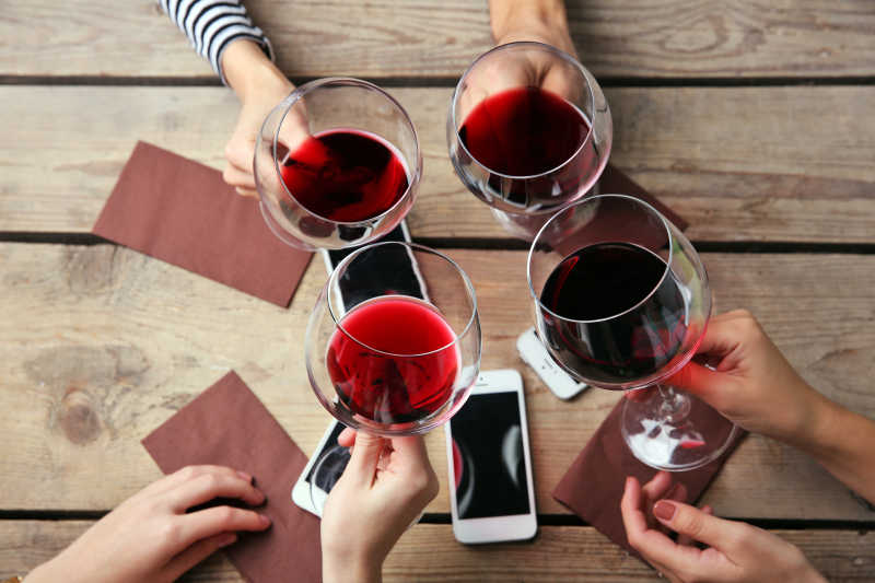木质桌板上的4只智能手机和拿着红酒杯干杯的人
