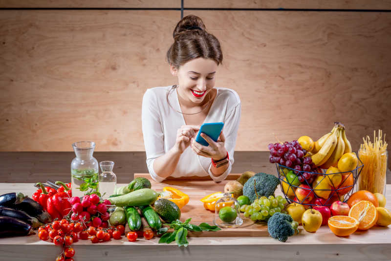 看手机的美女前面放着新鲜的水果和蔬菜