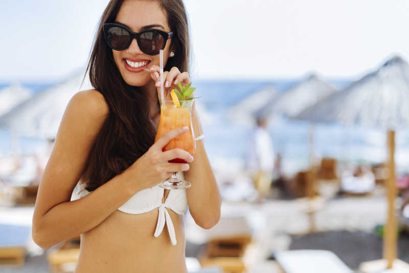 漂亮的黑发美女在沙滩上喝鸡尾酒享受着她的假期