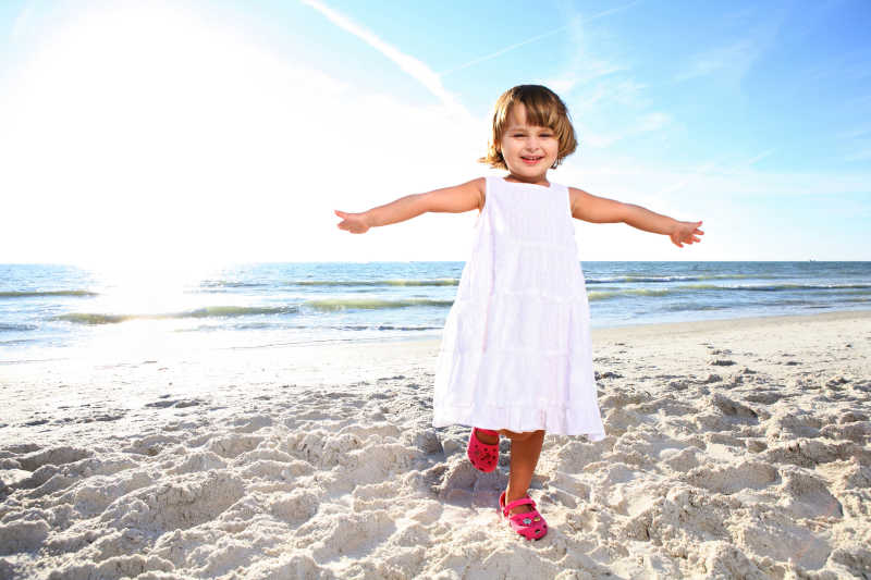 穿着白色连衣裙的小快乐女孩在海滩享受晴朗的日子向太阳射击