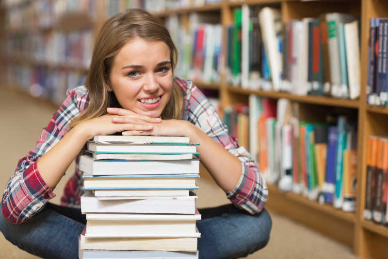 坐在图书馆地板上靠着一摞书的微笑的学生