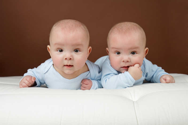 穿着蓝色衣服趴在床上看镜头的双胞胎宝宝