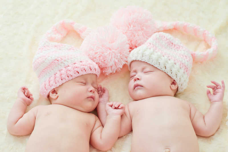 躺在白色毛毯上戴着粉色帽子睡着的双胞胎宝宝