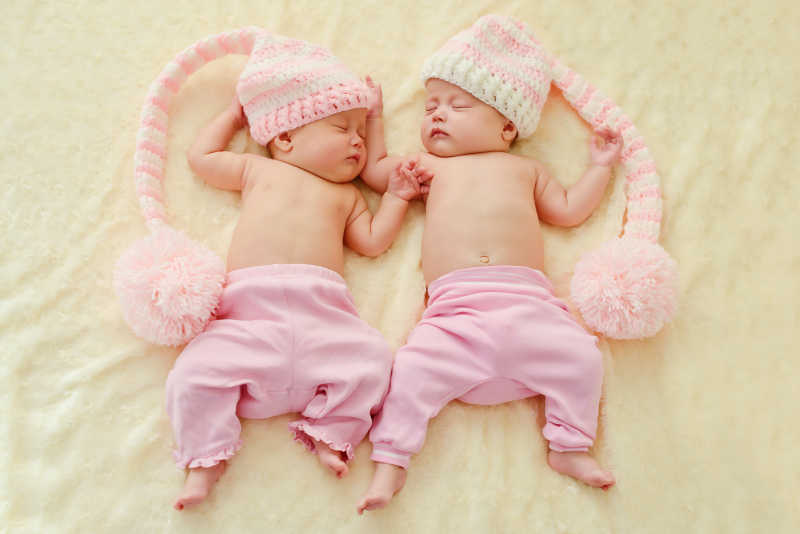 躺在米色床上穿戴着粉色衣帽睡着的双胞胎宝宝