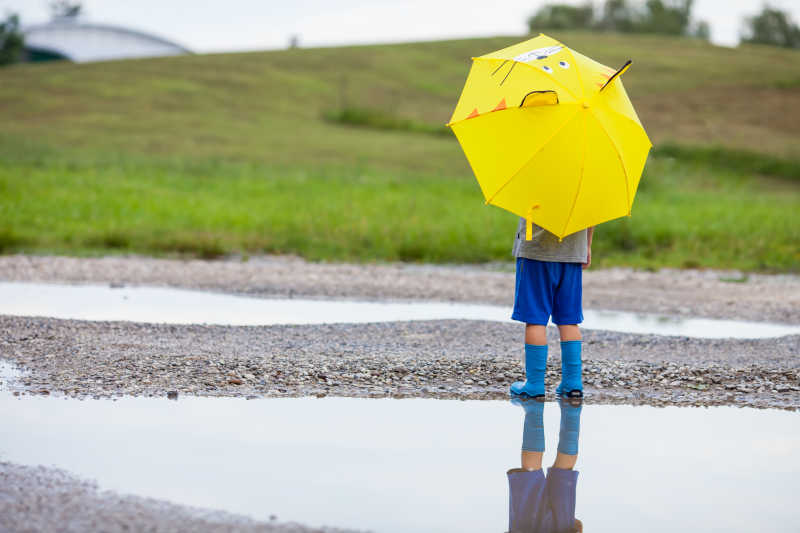 小男孩拿着黄色雨伞站在水坑旁边