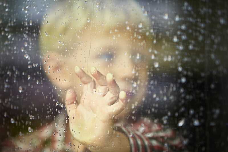 站在淋有雨水的窗户旁边的小男孩