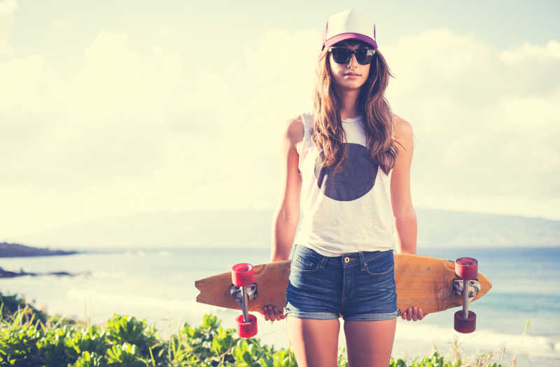 戴着墨镜和帽子手拿滑板的女子在海边