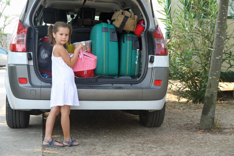 穿着白色裙子的小女孩把行李放在汽车的后备箱上准备度假