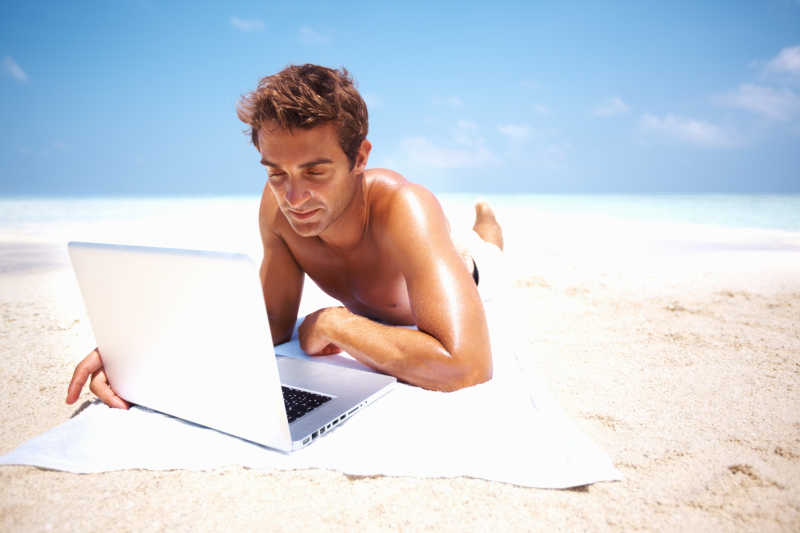 微笑啊的金发碧眼的男人趴在沙滩上使用笔记本电脑