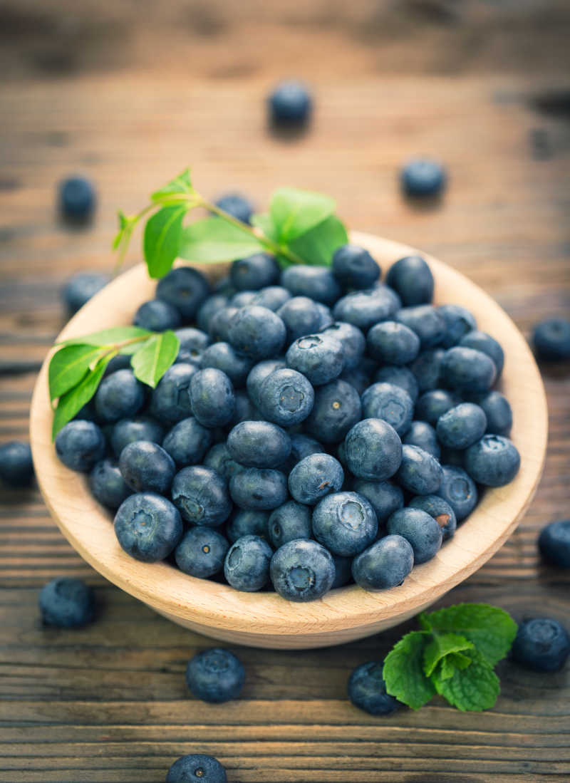 木桌上散落的蓝莓和碗中带着枝叶的蓝莓