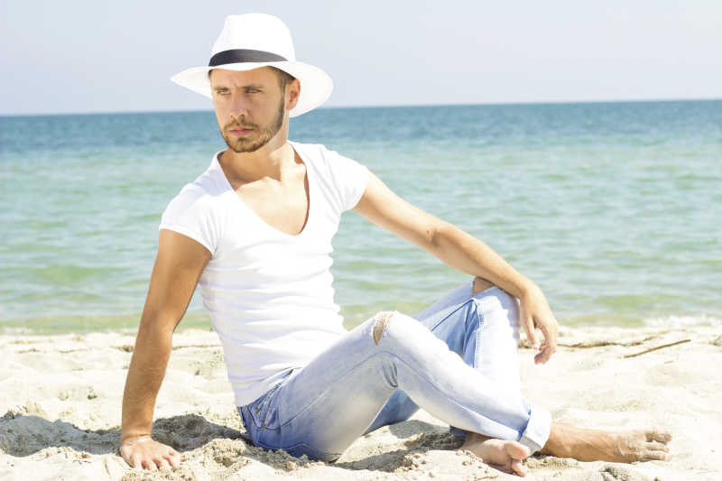 穿着白t和牛仔裤的年轻男人坐在沙滩上