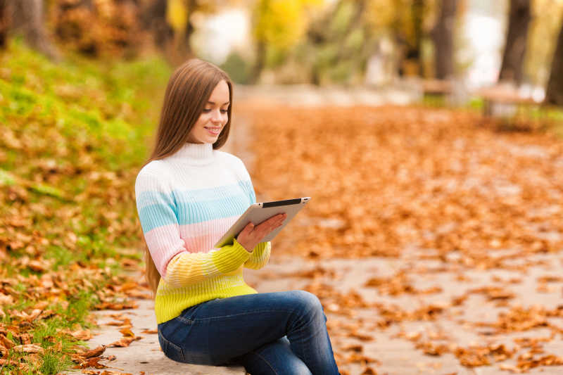 穿着毛衣和牛仔裤的年轻女孩坐在秋天的公园里看平板电脑