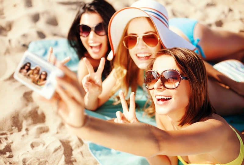 三个戴着太阳镜的漂亮的女孩在海滩上用手机自拍
