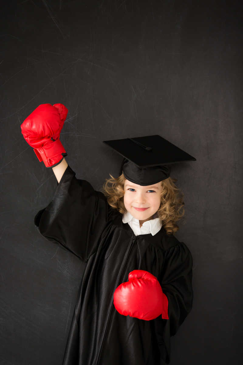 灰色背景下穿着学士服的学生戴着红色拳击手套