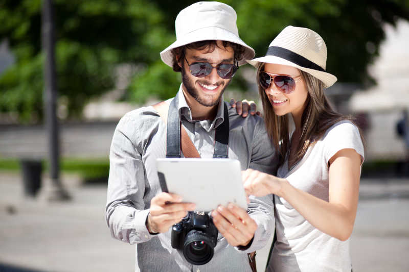 阳光下戴着帽子和墨镜使用平板电脑的年轻夫妇
