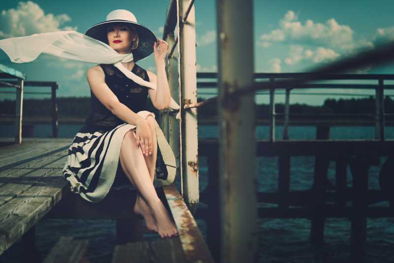 戴着白色帽子和红色衣服的时髦女人坐在老木头码头上