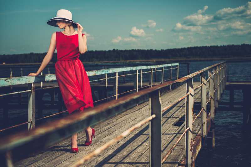 白色帽子和红色裙子的时髦女人站在老木头码头上