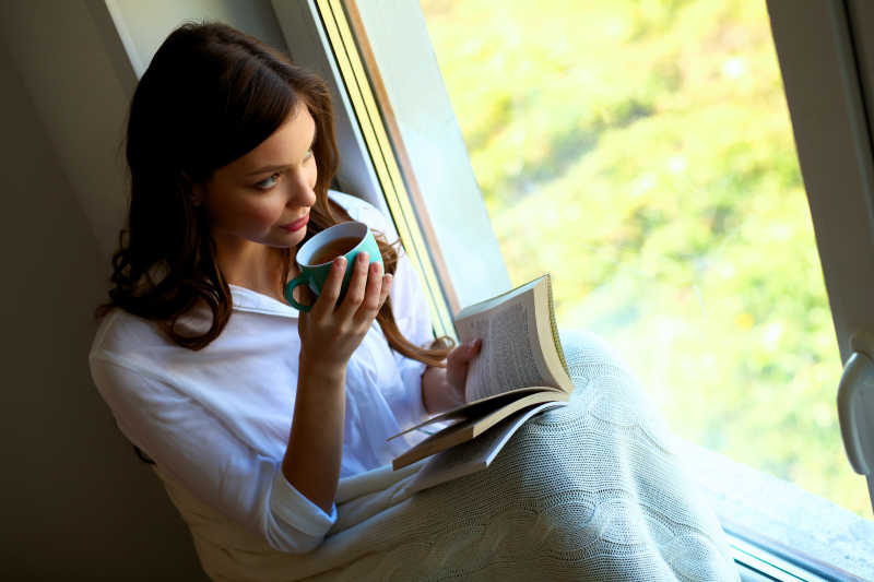 年轻女子坐在窗边看书和喝咖啡