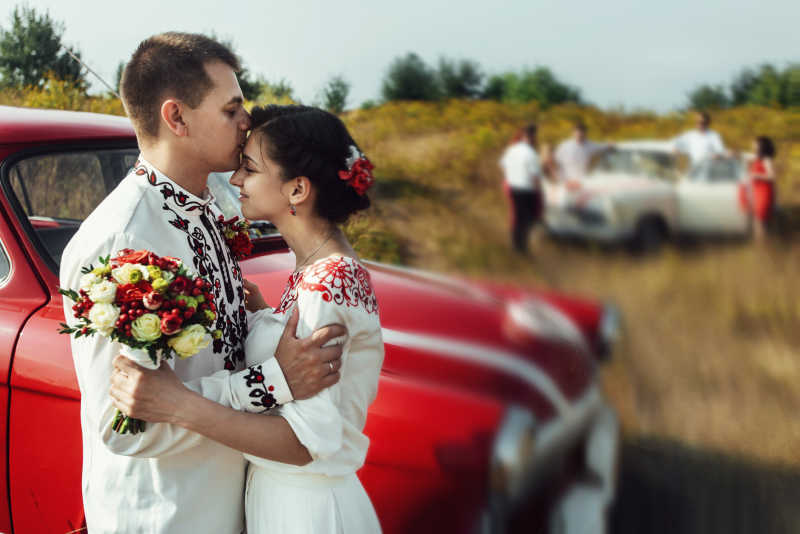 红色汽车旁边的新郎在亲吻新娘的额头