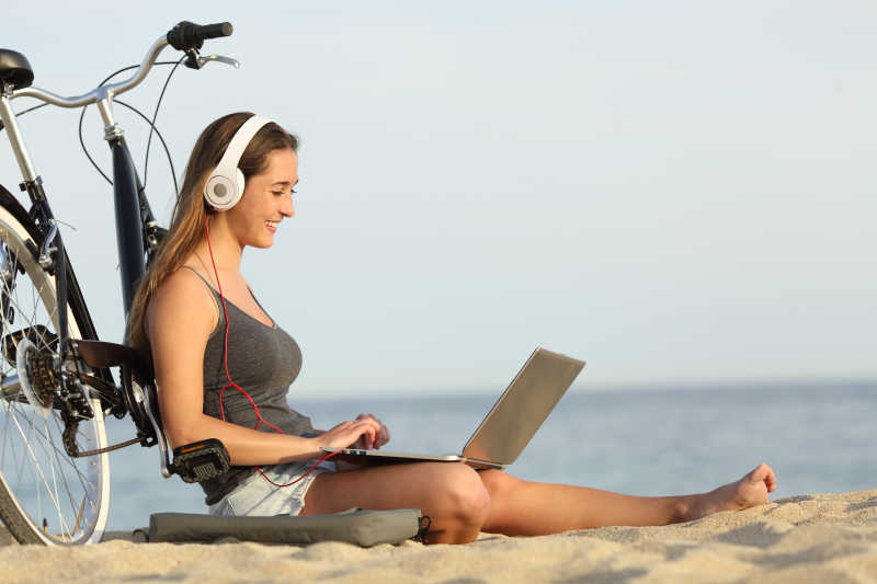 在海滩上靠着自行车用笔记本电脑学习的女孩