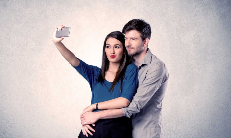 一对年轻夫妇在一个空的清灰墙背景概念前拿着手机自拍