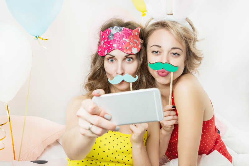 两个年轻的女孩拿着假胡子用手机拍照