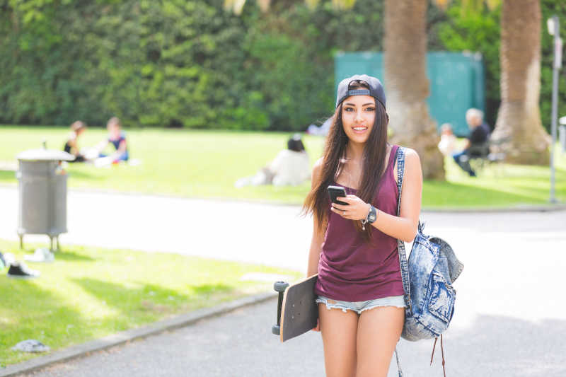 美丽的滑板女孩在公园里看智能手机