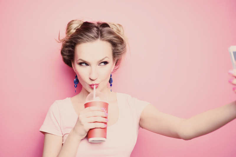 一个年轻漂亮的金发女孩在粉色背景下拿手机自拍用吸管喝饮料