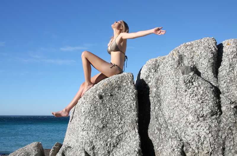 坐在岩石上张开双手享受阳光的比基尼美女