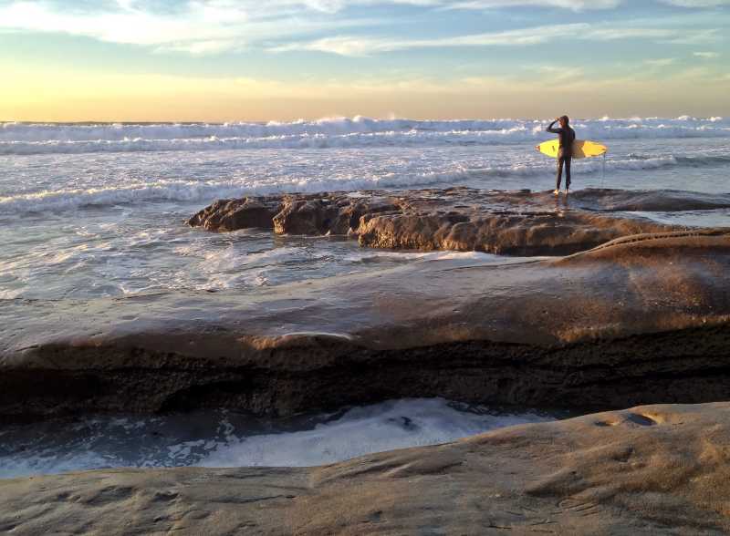 日出时分冲浪者站在岩石上看着海边