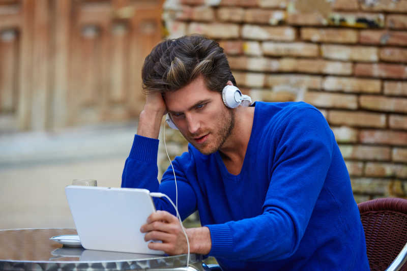 坐在户外咖啡馆桌前带着耳机看平板电脑的男人