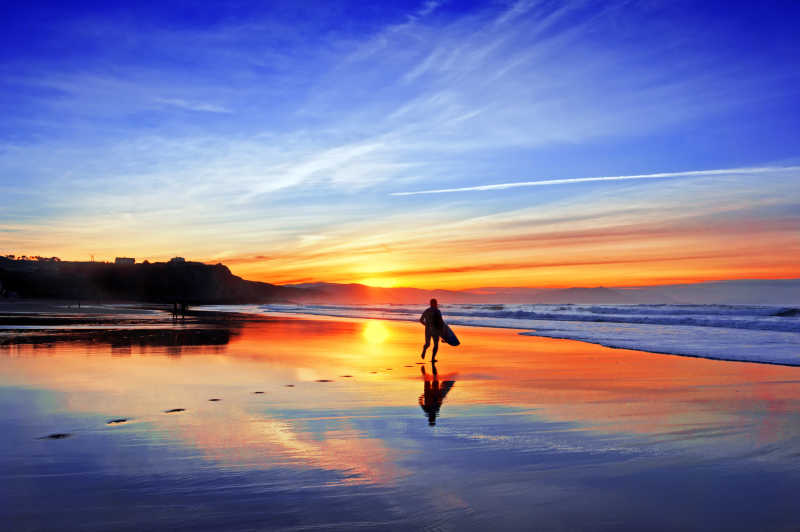 蓝天下日落时分拿着冲浪板走在海滩上的人