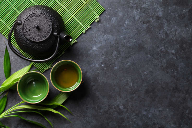 黑色桌面上绿色竹帘上的黑色茶壶和绿色茶杯