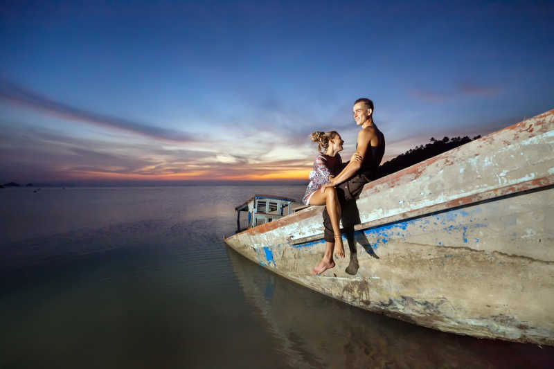 落日背景下一对年轻的夫妇笑着坐在沉船上