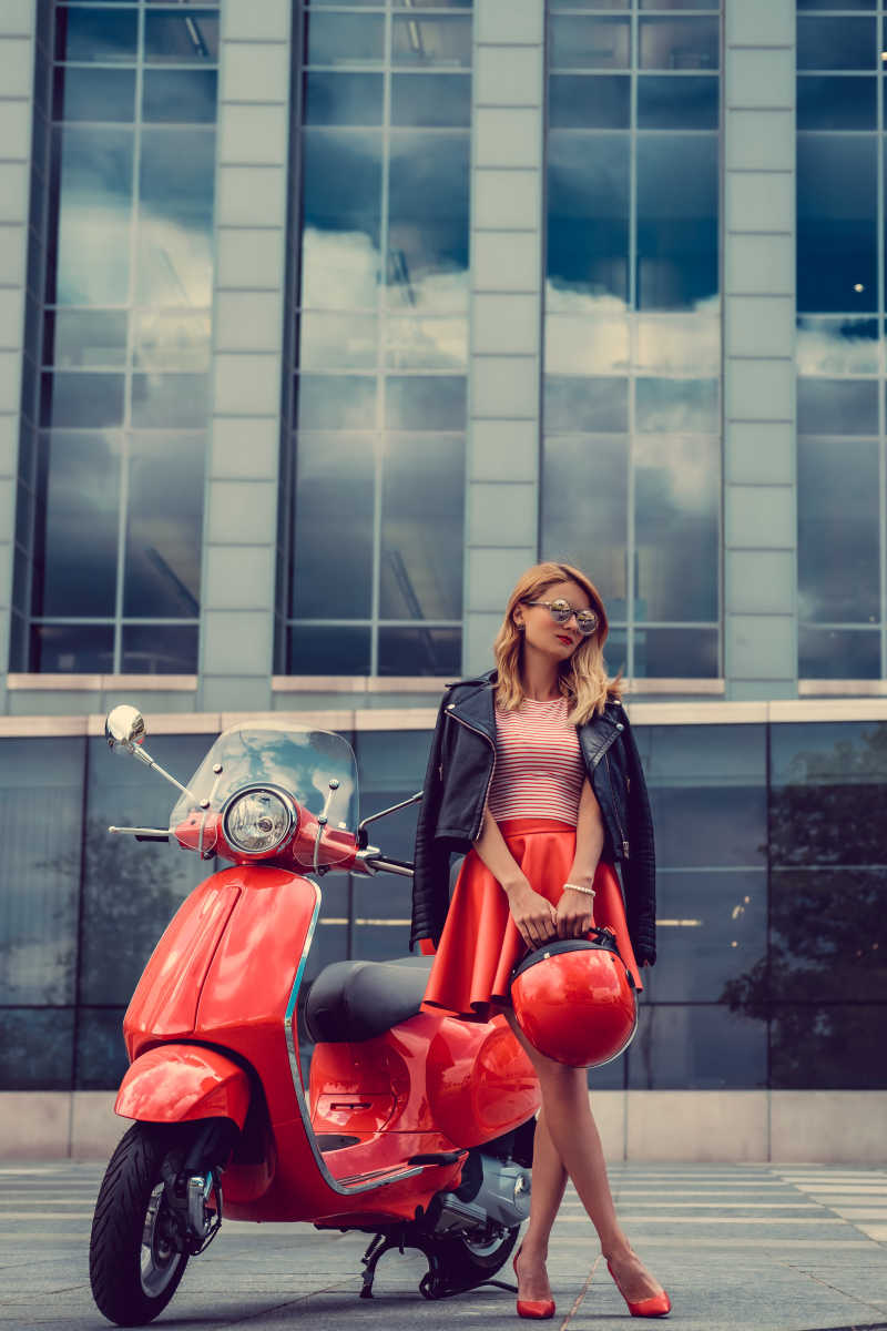 大厦前穿着红色裙子拿着头盔的美女站在电瓶车旁