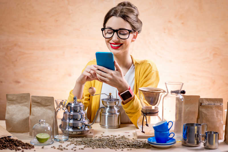 在摆满咖啡豆和咖啡用具的木桌前使用智能手机的美女
