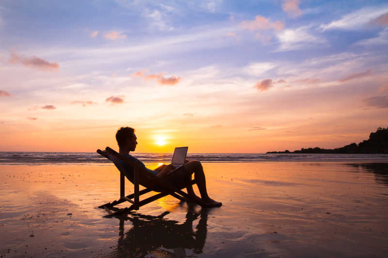 夕阳下海滩休闲椅上正用笔记本办公的男人