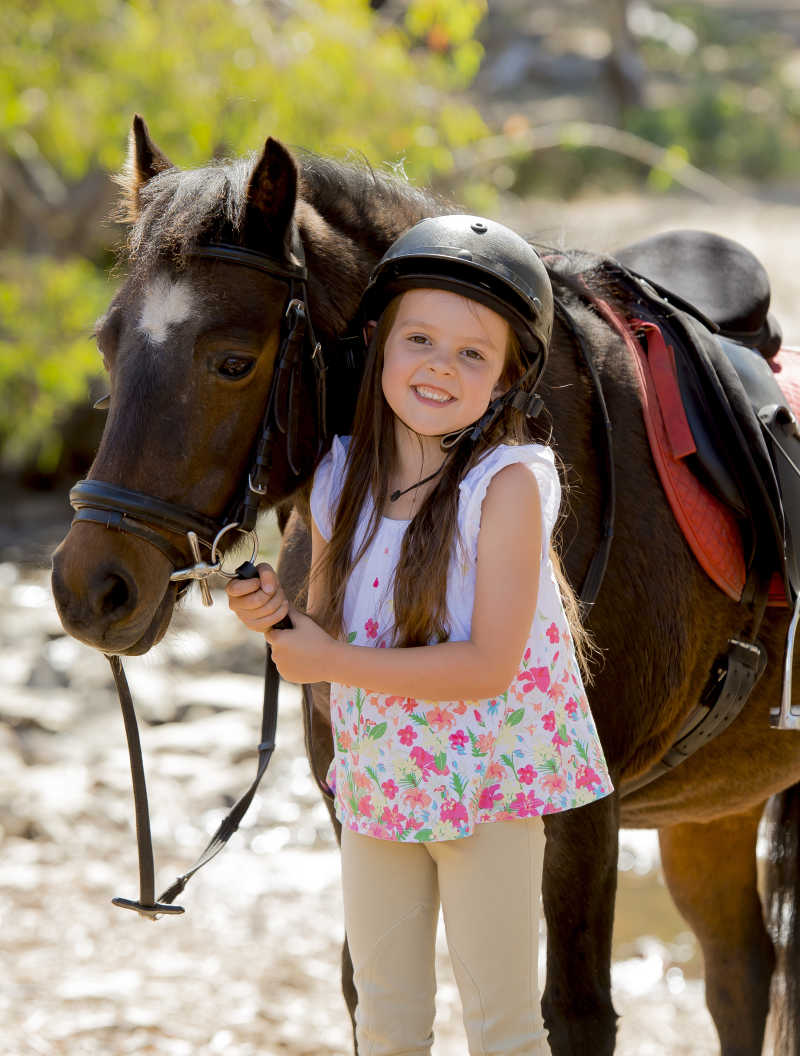 在户外带着头盔的小女孩牵着棕色的小马