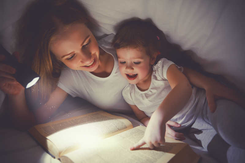 妈妈和女儿在床上用手电筒在毯子下看书