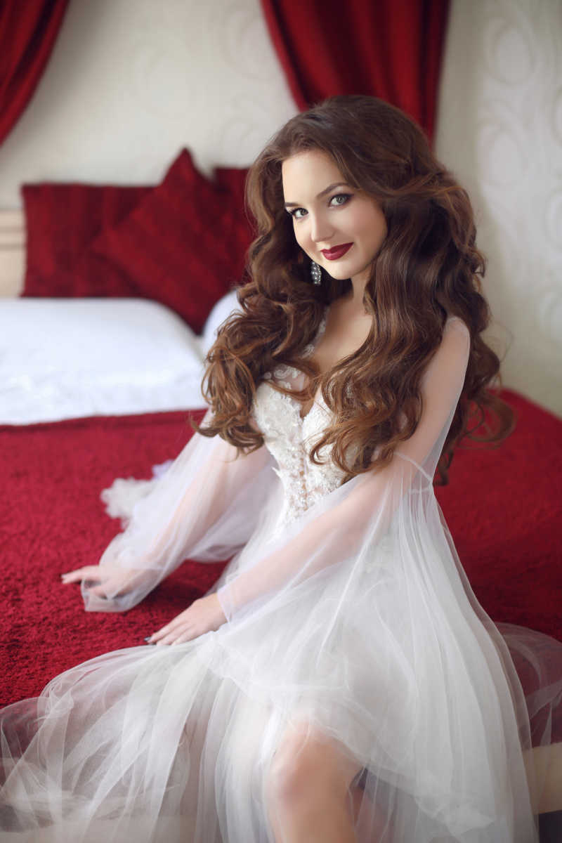 在床上穿着白色性感闺房礼服长袍的美丽微笑的卷发性感婚礼肖像