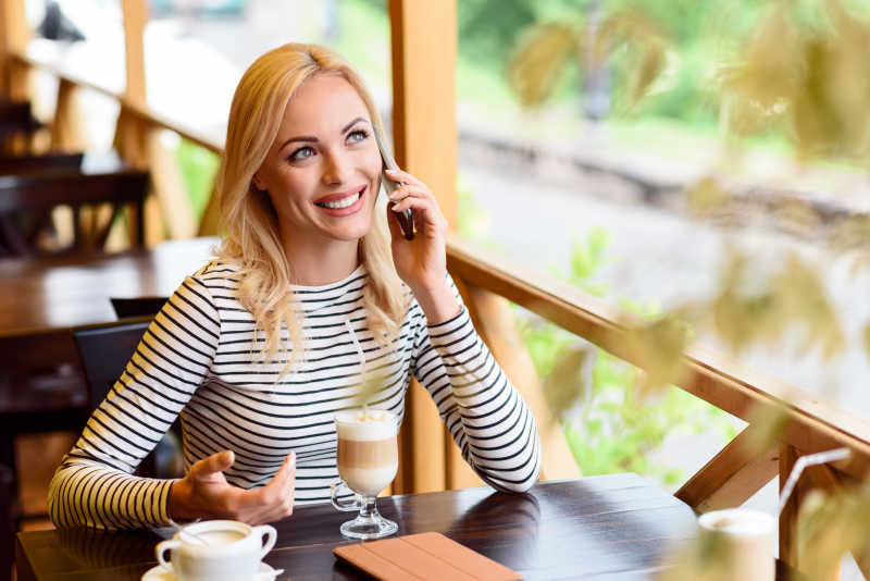坐在咖啡馆的桌子旁用电话交流的漂亮的金发女郎