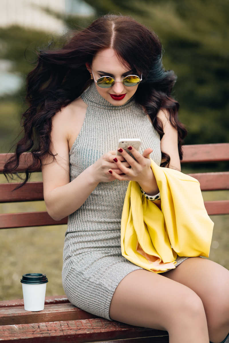 时尚性感的女人坐在坐在街上的长凳上使用智能手机